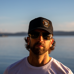 Man with sunglasses wearing a Black 'Hunt Fish Maniac' Logo Trucker Hat at the dock.