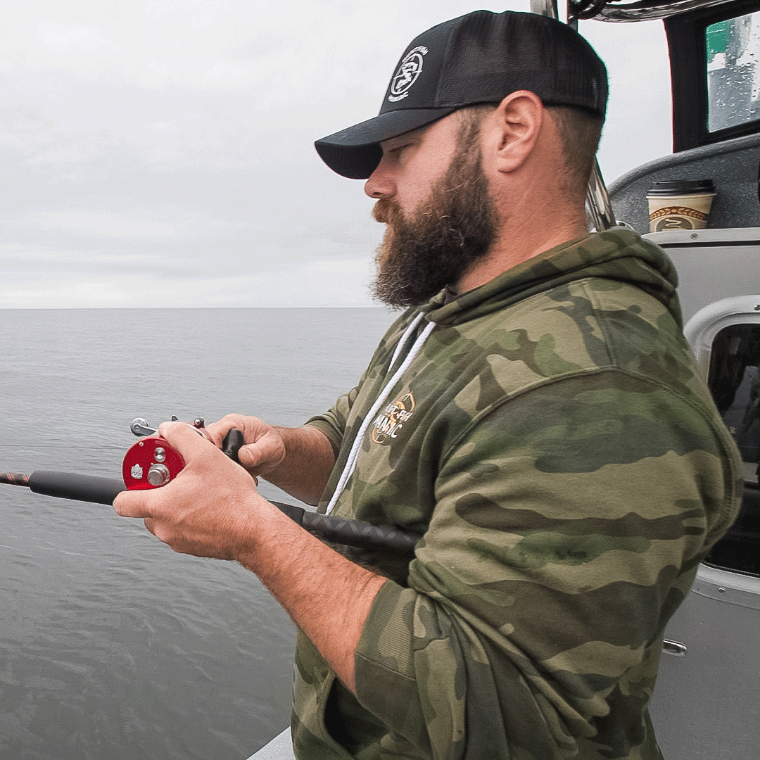 Man with a beard wearing Camo 'Hunt Fish Maniac' Pullover Hooded Sweatshirt while fishing on a boat.