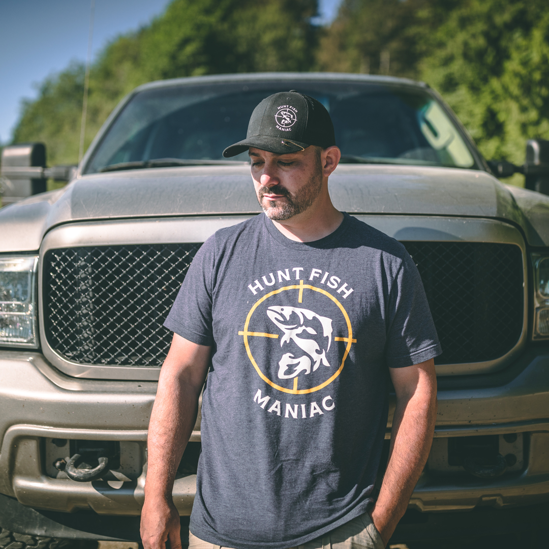 Paul Marier standing in front of a truck wearing a Heather Navy 'Hunt Fish Maniac' Logo Crewneck Tee.