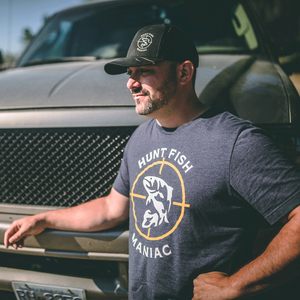 A man leaning against a truck wearing a Heather Navy 'Hunt Fish Maniac' Logo Crewneck Tee and black hat.