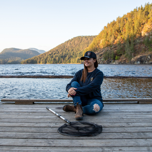 A woman sitting on a dock wearing a Heather Black 'Hunt Fish Maniac' Logo Long-Sleeve Tee and black hat.