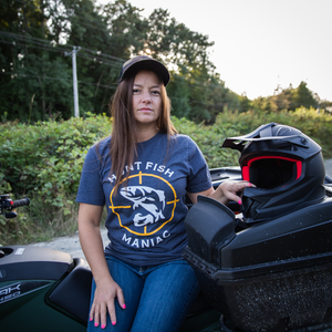 A woman siitting on a quade holding a helmet wearing a Heather Navy 'Hunt Fish Maniac' Logo Crewneck Tee.