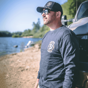 Paul Marier leaning against a truck wearing a Heather Black 'Hunt Fish Maniac' Logo Long-Sleeve Tee, black hat and sunglasses.