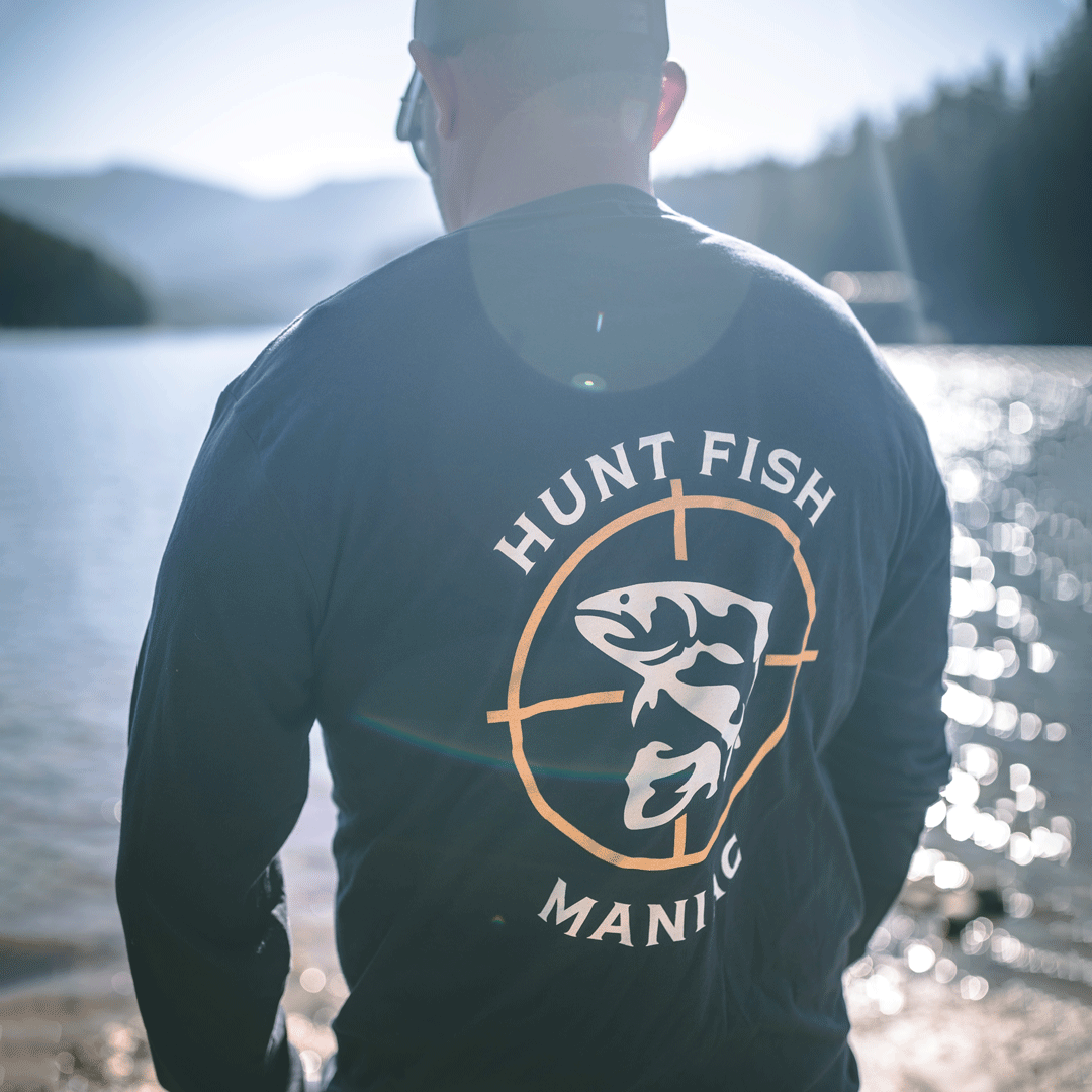 Man standing by a river wearing a Heather Black 'Hunt Fish Maniac' Logo Long-Sleeve Tee.