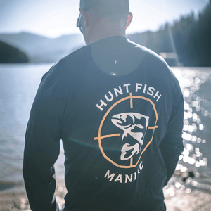 Man standing by a river wearing a Heather Black 'Hunt Fish Maniac' Logo Long-Sleeve Tee.