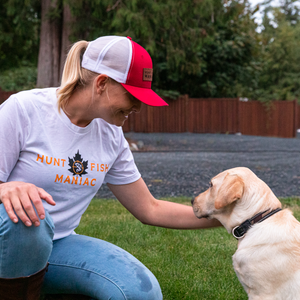 A woman petting a puppy, wearing a Red & White 'Hunt Fish Maniac' Leather Banner Logo Baseball Hat and white tee shirt.