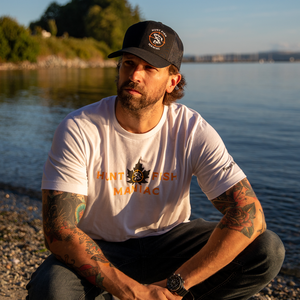 A man sitting by the shore at sunset wearing a White 'Hunt Fish Maniac' Classic Maple Leaf Crewneck Tee and black hat.